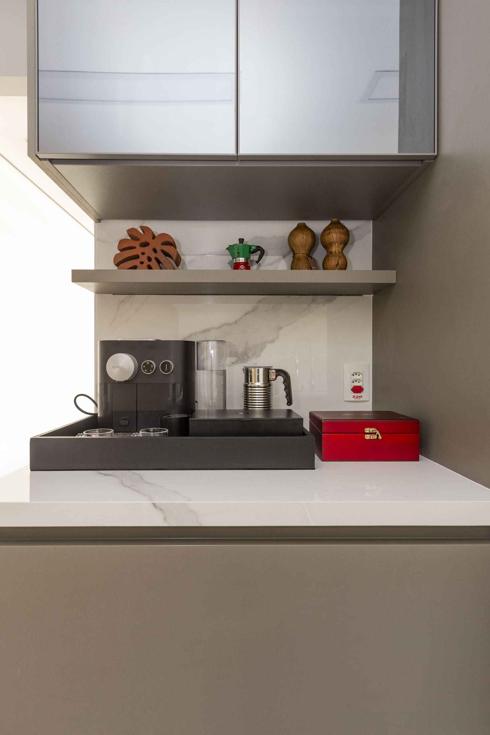Canto do café elegante com cafeteira preta, espumador de leite inox e caixa vermelha sobre bancada branca brilhante. Prateleira cinza com itens decorativos como moedores de madeira, mini cafeteira italiana e adorno de cerâmica, com fundo em mármore branco.