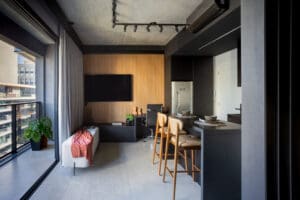 Ambiente de apartamento moderno com decoração industrial, integrado entre sala de estar, cozinha e varanda envidraçada, com bancada preta, cadeiras altas de madeira e um painel de TV em madeira.