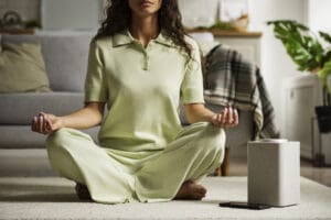 Mulher sentada em posição de meditação no chão de uma sala, com as pernas cruzadas e as mãos apoiadas nos joelhos em gesto de mindfulness, ao lado de uma caixa de som inteligente e um smartphone.